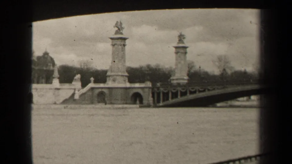 1947 Paris frame showing the river foreground and a monumental bridge approach with sculptural pillars and bare trees behind.