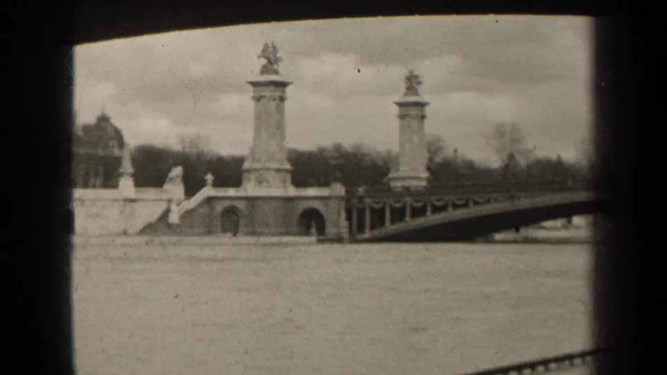 1947 Paris home-movie frame showing a grand stone bridge entrance with twin pylons, statues, railings, river, and cloudy sky.