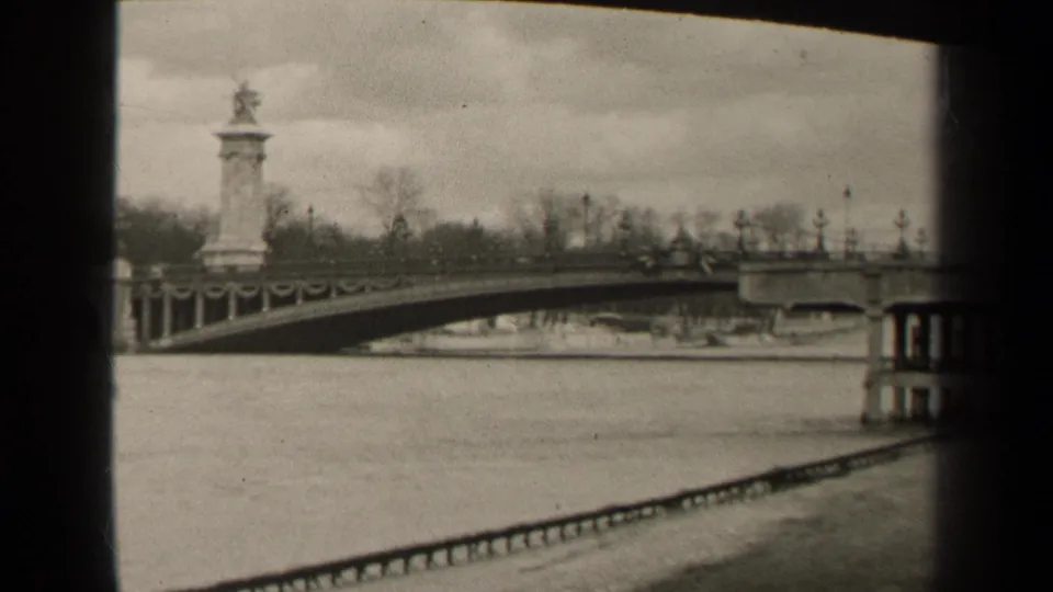 1947 Paris frame showing the bridge span and railing across the river, a stone pylon, bare trees, cloudy sky, and black film-frame edge.