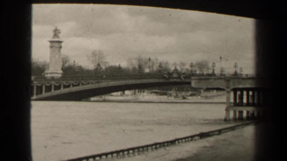 1947 Paris home-movie frame showing a river, arched bridge, bare trees, and a small car crossing under an overcast sky.