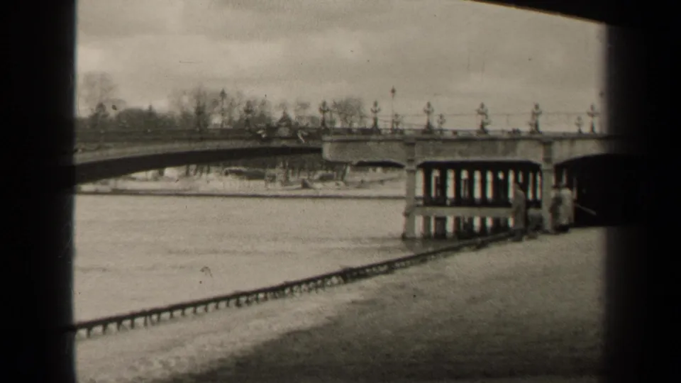1947 Paris home-movie frame with a river bridge, embankment railing, bare trees, and small pedestrians near the right support.