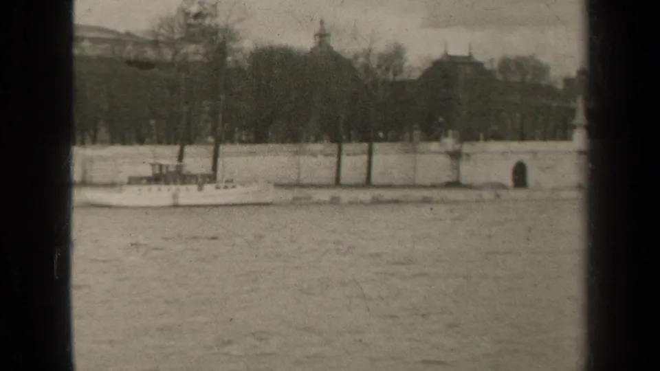 1947 Paris riverfront with a small boat near the embankment, winter trees, stone wall, and historic buildings across the water.