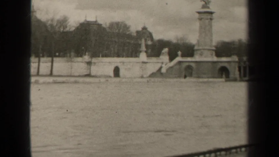 1947 Paris home-movie frame showing a broad river crossing with ornate bridge columns, stone arches, bare trees, and distant buildings.
