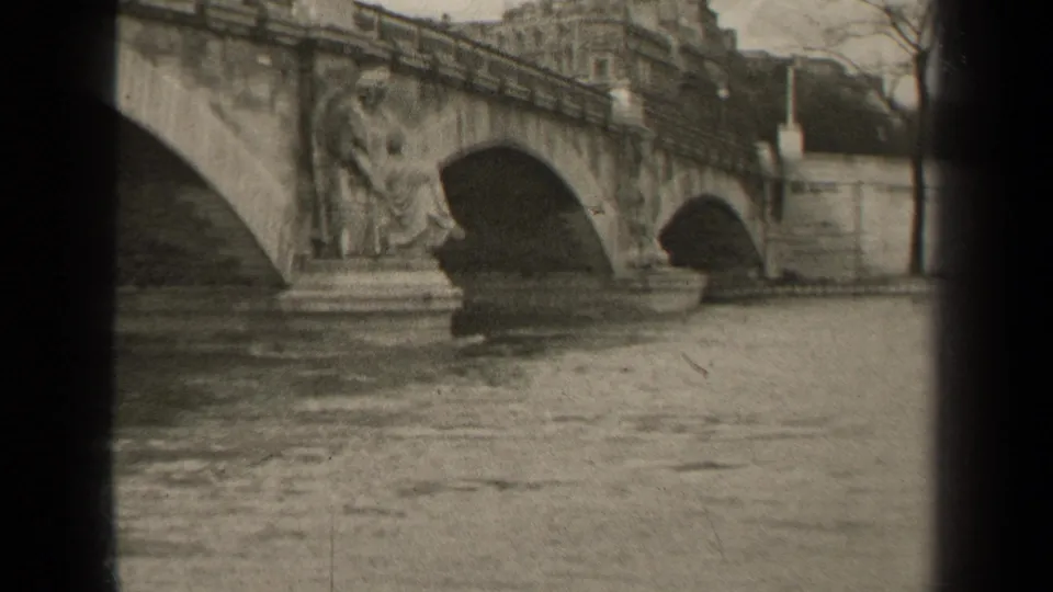 1947 Paris home-movie frame showing a stone bridge with arched spans, sculpted figures, river water, and buildings beyond.