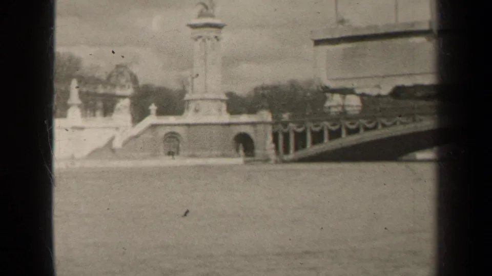 1947 Paris home-movie frame showing a riverside monument, bridge approach, stone balustrade, river water, and pale overcast sky.