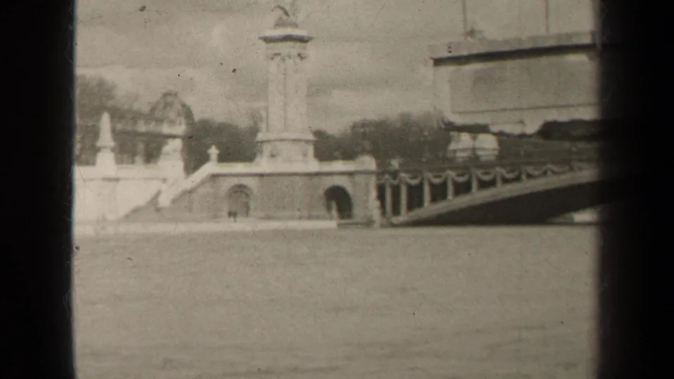 1947 Paris home-movie frame showing a tall stone monument, bridge railings, and riverside architecture in soft black-and-white 8mm.