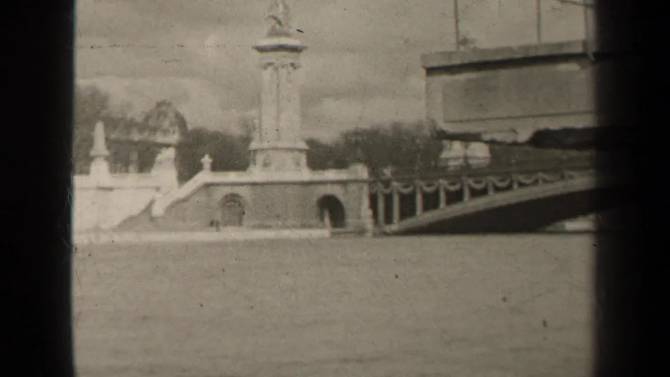 Black-and-white 8mm view of a Paris bridge end, column monument, ornate railing, and river surface, partly framed by dark film edges.