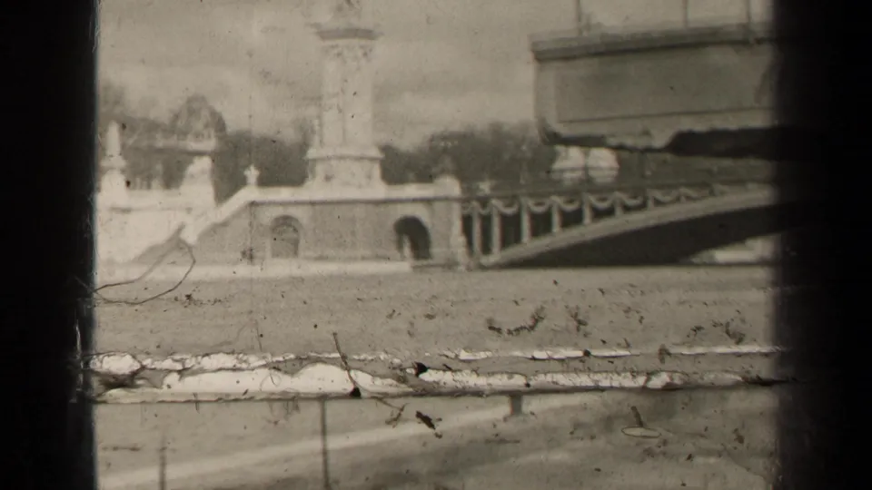 1947 Paris frame with a bridge approach, tall monument, river foreground, and pronounced scratches and exposure variation from the 8mm film source.