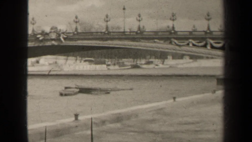 1947 Paris home-movie frame showing an ornate bridge railing, lamps, riverboats and paved embankment in black and white.