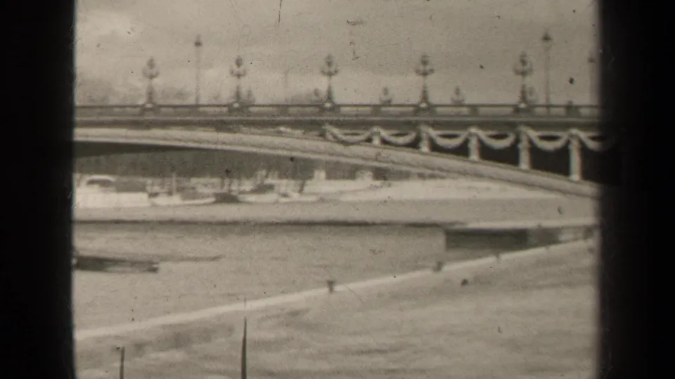 Black-and-white 1947 Paris frame with bridge lamps and banners above the river, with a boat near the embankment.