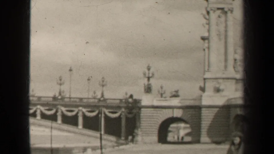 1947 Paris home-movie frame showing a stone bridge approach with arched opening, ornate lampposts, draped decoration, and part of a monument.