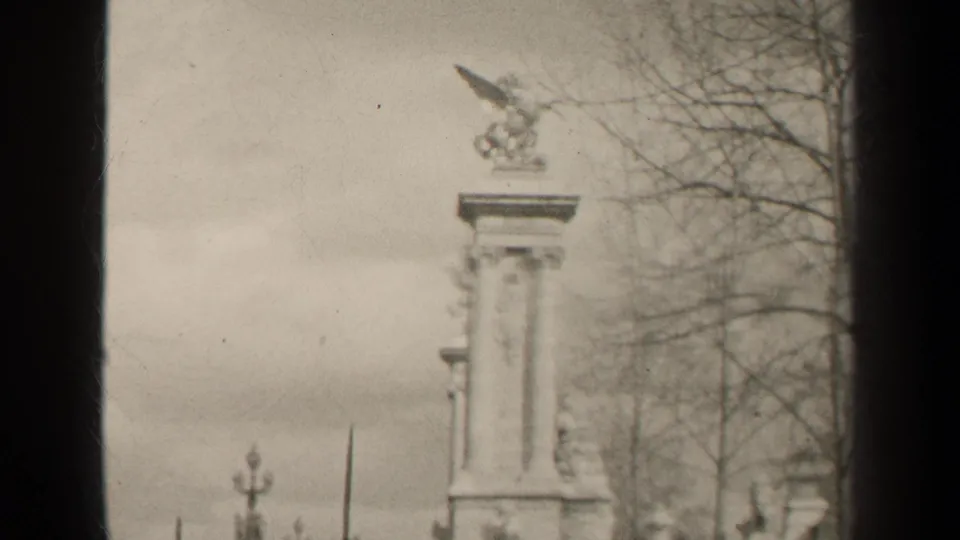 1947 Paris home-movie view of the monument pedestal and winged horse sculpture framed by bare branches and soft overcast light.