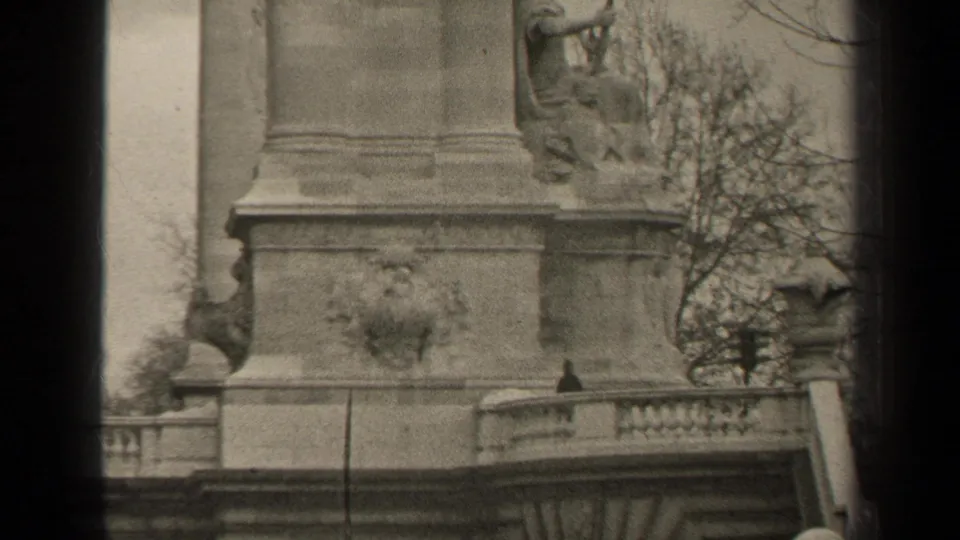 1947 Paris home-movie frame showing a stone monument pedestal, seated sculpture, railing, and bare trees in soft black-and-white grain.