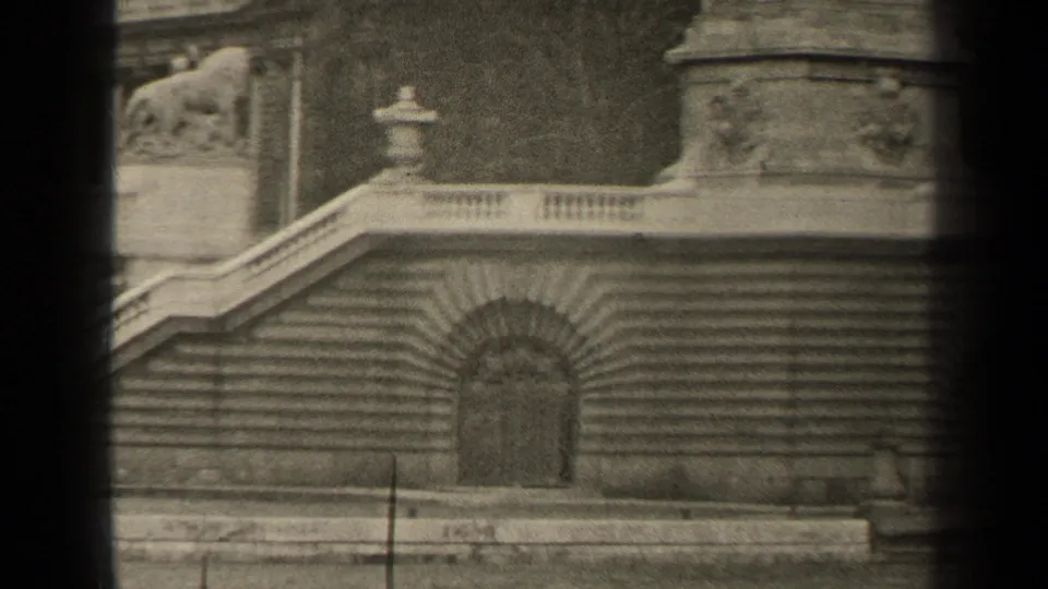 1947 Paris home-movie frame showing lower monument stairs, a recessed arched doorway, balustrade, and lion sculpture above.