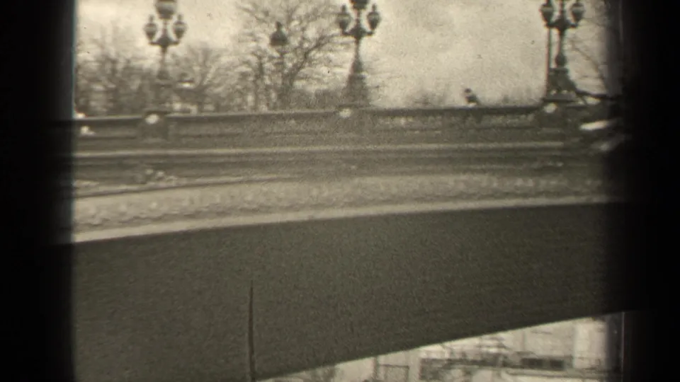 1947 Paris home-movie frame showing a decorated bridge balustrade, globe lampposts, bare trees, and passing-vehicle motion blur.
