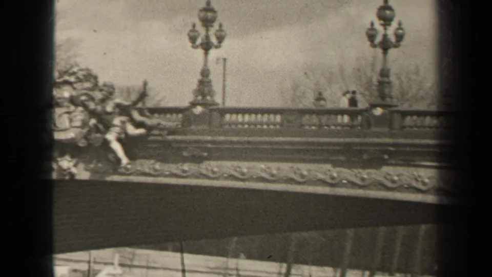 1947 Paris frame showing an ornate bridge railing, globe lampposts, partial sculpture, bare trees, and distant pedestrians along the walkway.
