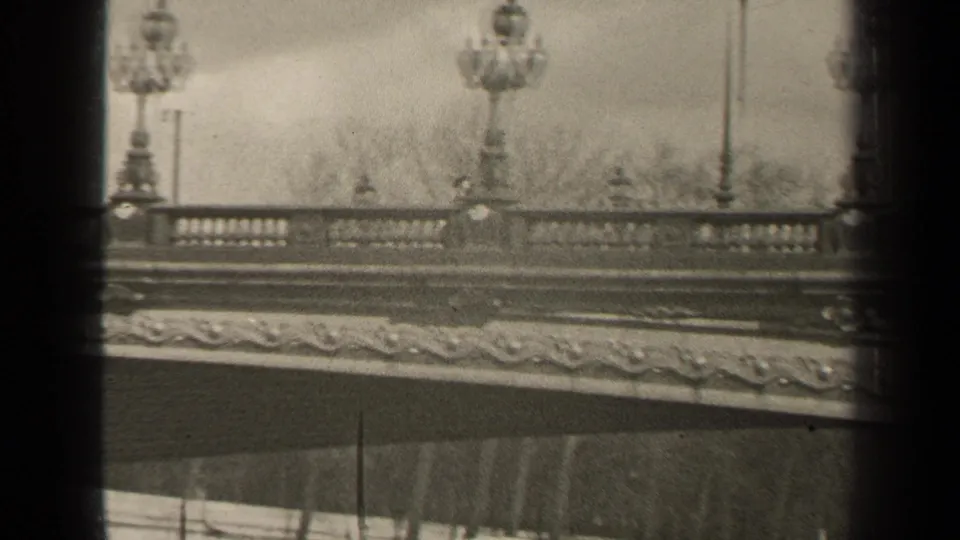 1947 Paris home-movie frame showing an ornate bridge railing, classical lampposts, and soft black-and-white 8mm texture.