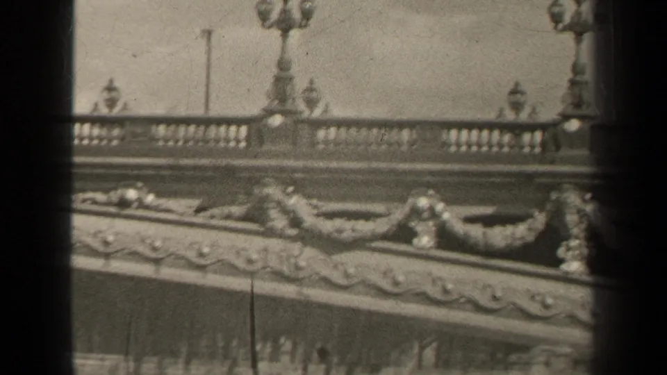 Black-and-white 8mm frame from Paris showing carved bridge garlands, balustrade, and lamp standards during a moving pan.