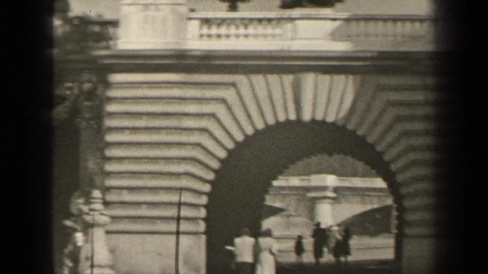 1947 Paris home-movie frame showing several pedestrians moving near a large stone arch with heavy foreground shadows.