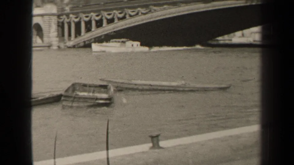 Black-and-white 8mm view of river craft beneath an ornate Paris bridge, with quay wall in front.