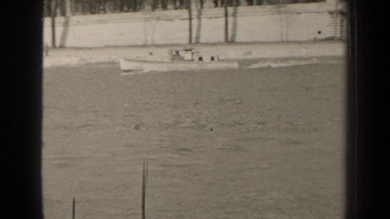 1947 Paris home-movie frame showing a small boat moving across calm water beside a pale embankment wall and bare trees.