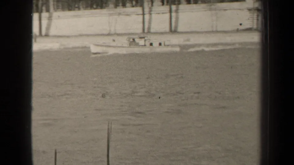 1947 Paris home-movie frame showing a small boat moving across calm water beside a pale embankment wall and bare trees.
