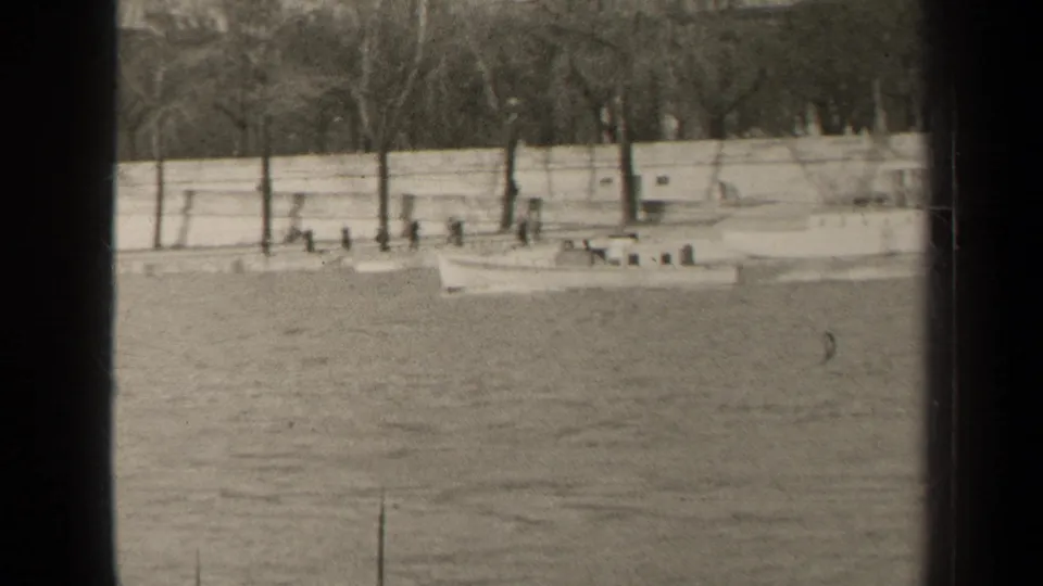 1947 Paris home-movie frame showing a small boat on rippled water beside a stone quay, bare trees, and distant pedestrians.