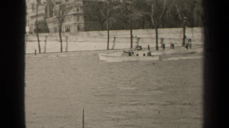 A small civilian boat travels along a Paris waterfront, with a pale stone embankment, bare trees, and apartments in the background.
