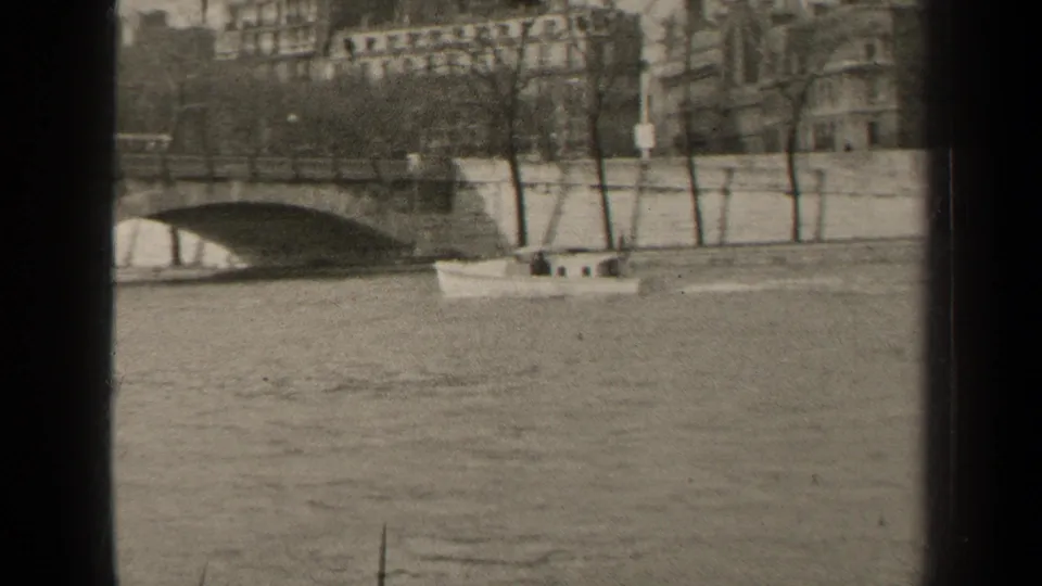 The boat moves beside a Paris embankment as a stone bridge enters the background, with pedestrians and buildings visible beyond.