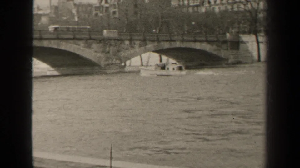 Black-and-white 8mm view of the Seine in Paris, with bridge arches, a small boat, bare trees, and riverside buildings.