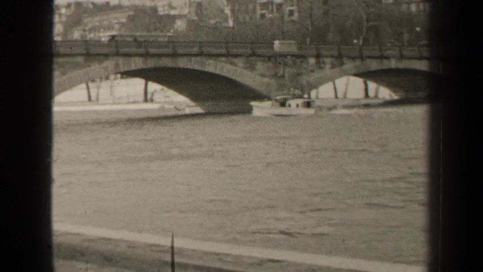 Paris Seine frame from 1947 showing a small boat by a stone arch bridge, with railings, bare trees, and soft 8mm grain.