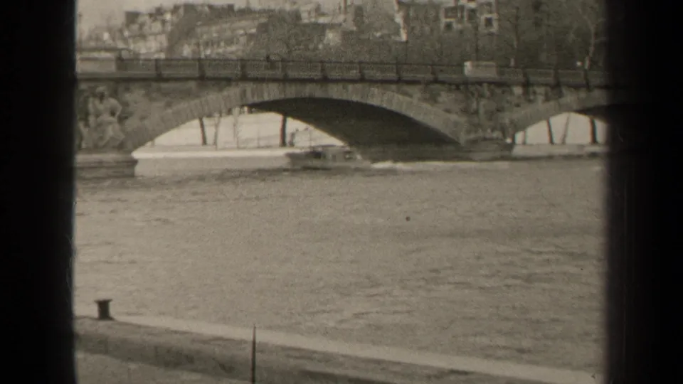 1947 Paris home-movie frame showing the Seine, a stone arch bridge, riverside wall, small boat, trees, and buildings.