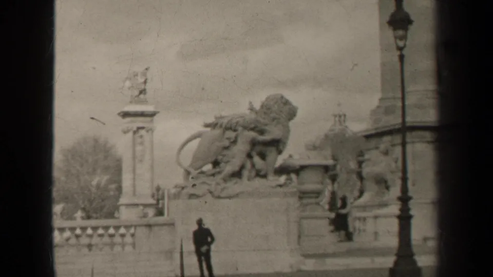 1947 Paris home-movie frame showing a stone monument with lion statuary, a balustrade, street lamp and a distant standing figure.
