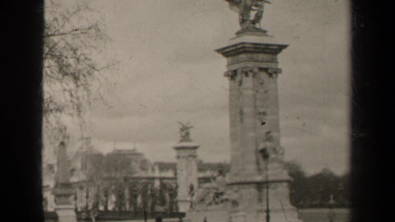 Tall stone column with sculpture, smaller monument, trees, railings and distant pedestrians.