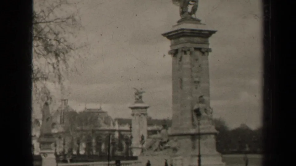 1947 Paris home-movie frame centered on a tall stone column beside bare trees, distant architecture, railings and pedestrians.