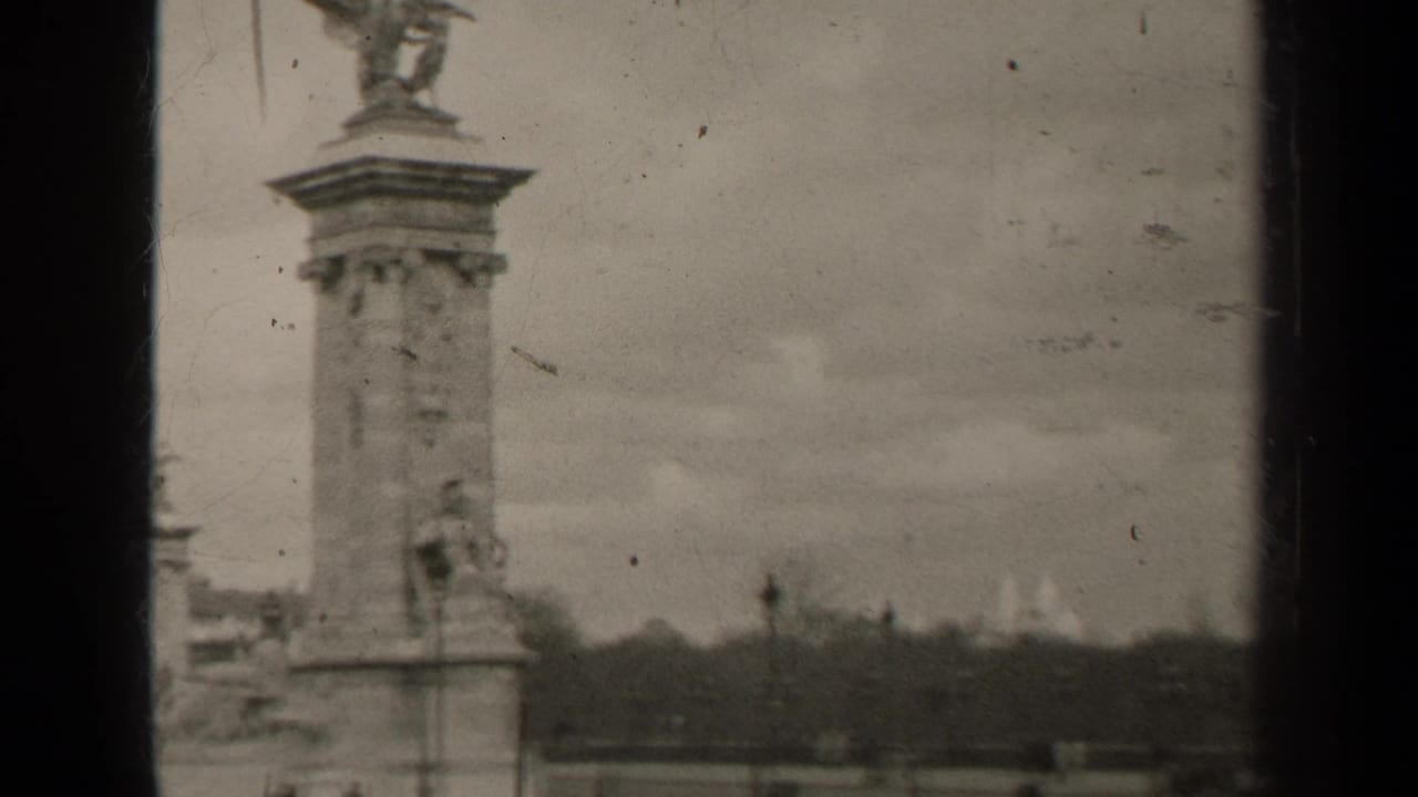 1947 Paris home-movie frame showing a Pont Alexandre III pylon with winged sculpture, soft focus, scratches, and cloudy sky.