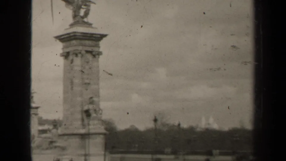 1947 Paris home-movie frame showing a Pont Alexandre III pylon with winged sculpture, soft focus, scratches, and cloudy sky.