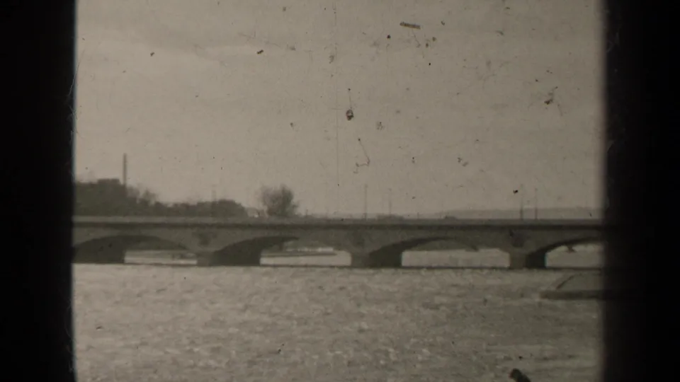 1947 Paris frame looking across the Seine toward bridge arches, embankment trees, and soft 8mm film scratches.