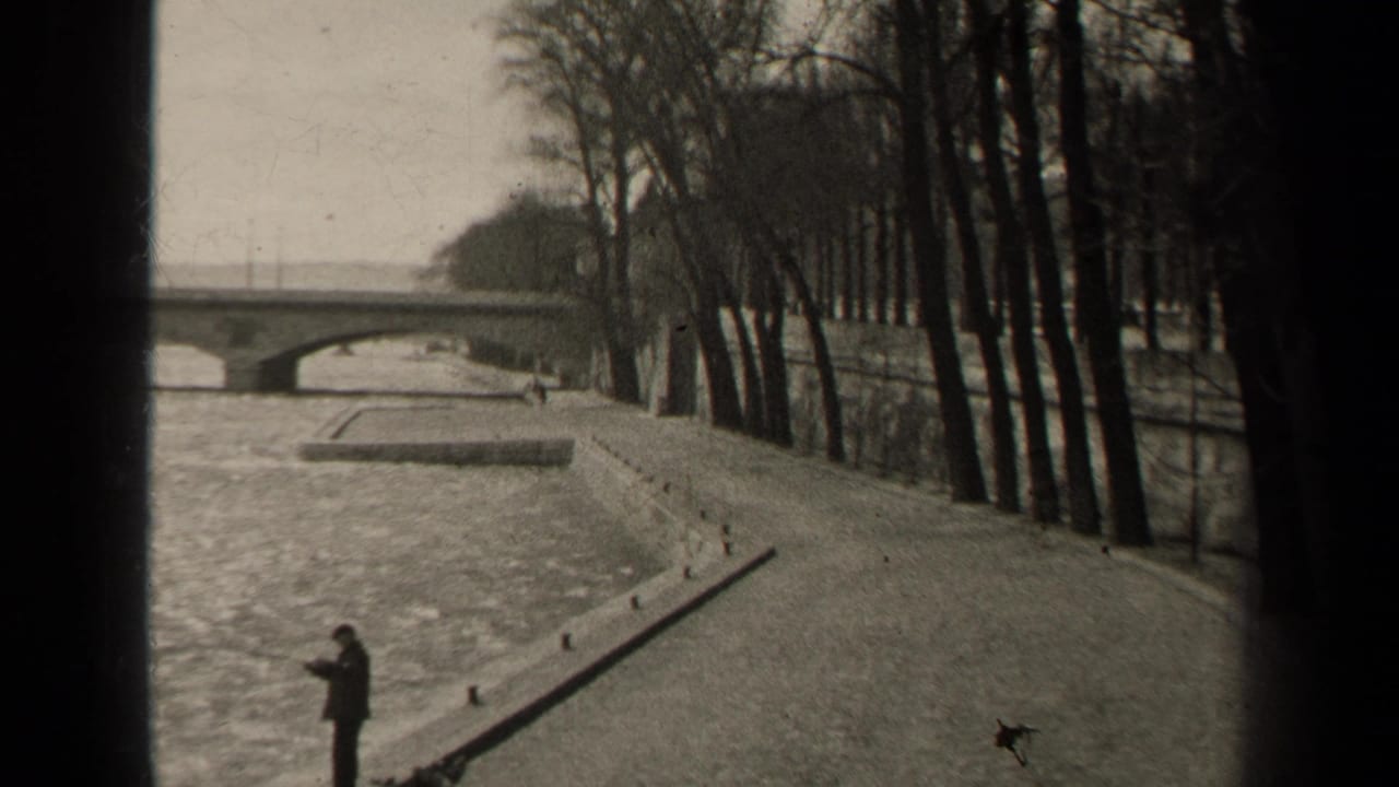 Man in dark clothing standing beside a riverbank path with a bridge and bare trees beyond.