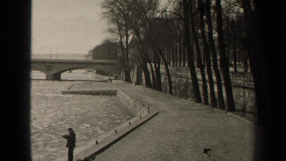 1947 Paris home-movie frame showing a solitary man by the river, a paved embankment, bare trees, and a bridge in the background.