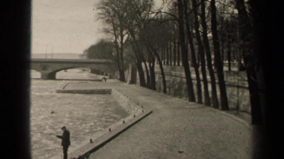 Black-and-white 8mm view of a Paris riverbank with a man near the water, leafless trees lining the path, and a bridge beyond.