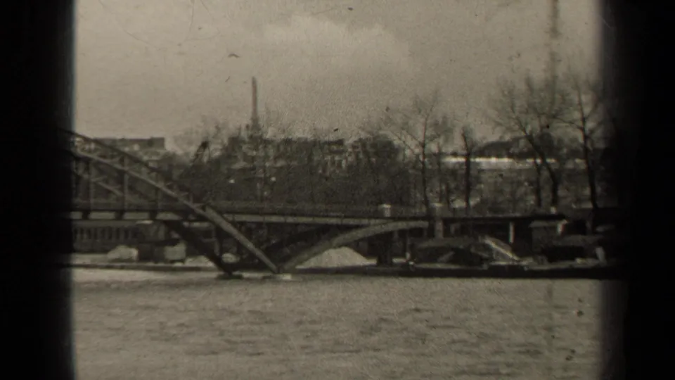Black-and-white Paris home-movie frame showing a bridge over the river, distant buildings, bare trees, and the Eiffel Tower in the background.