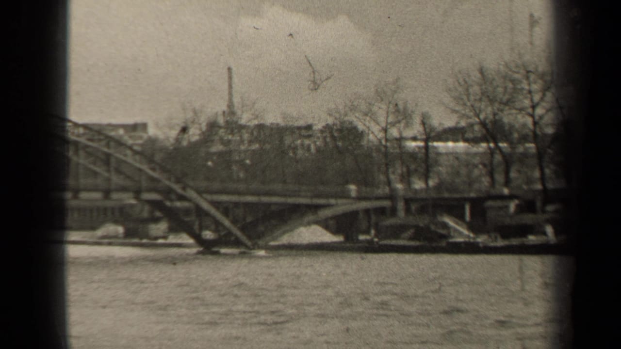 River water flowing beneath a steel bridge with bare trees and buildings beyond.