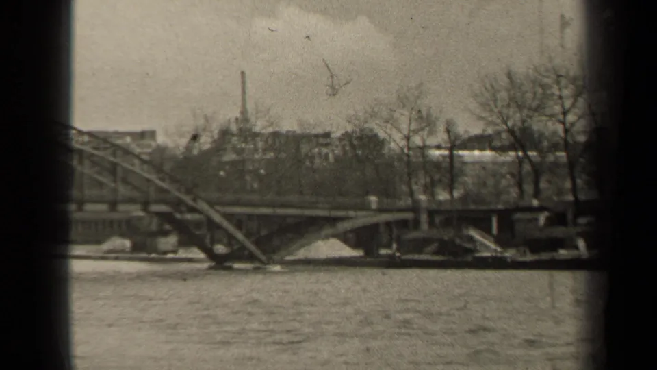 1947 Paris home-movie frame showing rippling river water, a steel bridge, bare riverside trees, and distant urban buildings.