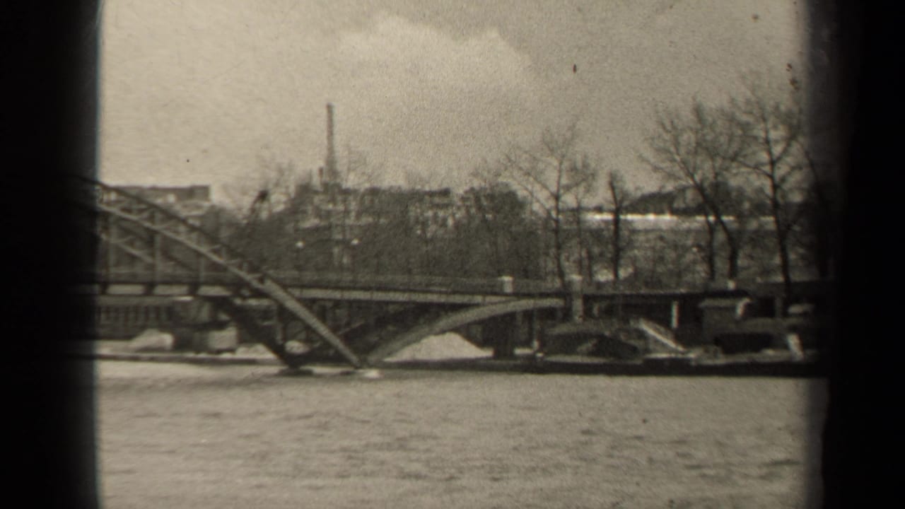 Steel bridge over a river with bare trees and low buildings in the background.