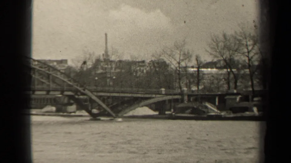 1947 Paris frame of a steel bridge crossing rippled river water, with leafless trees and urban buildings along the far bank.