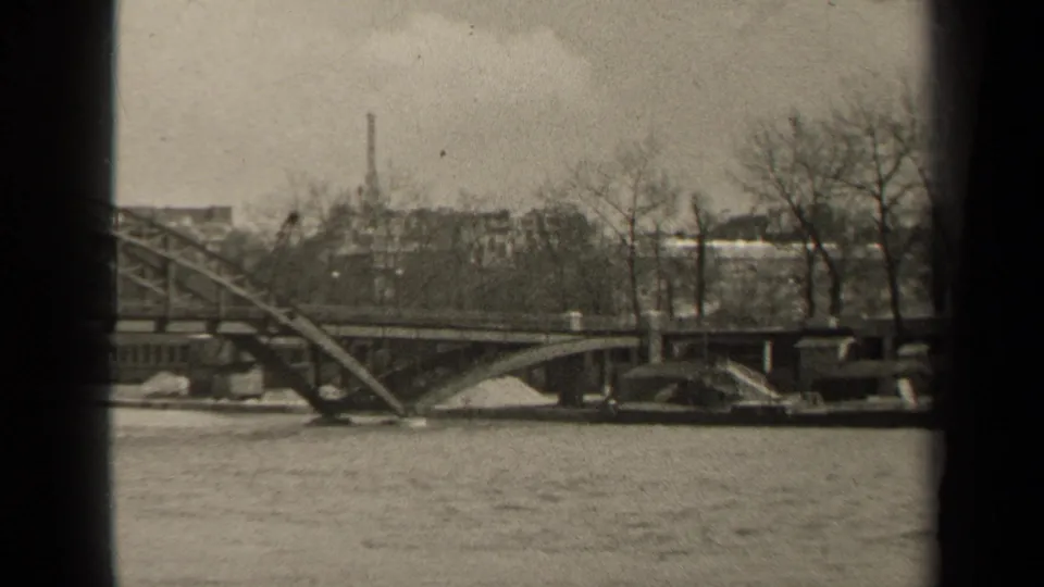 1947 Paris home-movie frame showing a steel bridge across a river, with leafless trees, older buildings, and dark film-edge masking.