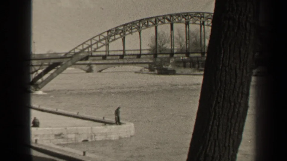 1947 Paris frame showing an arched steel bridge, stone river quay, bare trees, a large foreground tree trunk, and small distant pedestrians.