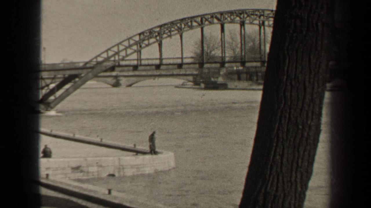 1947 Paris home-movie frame showing high river water beneath a steel bridge, with observers on a stone quay and dark tree trunks framing the view.