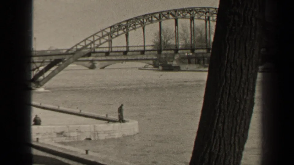 1947 Paris home-movie frame showing high river water beneath a steel bridge, with observers on a stone quay and dark tree trunks framing the view.